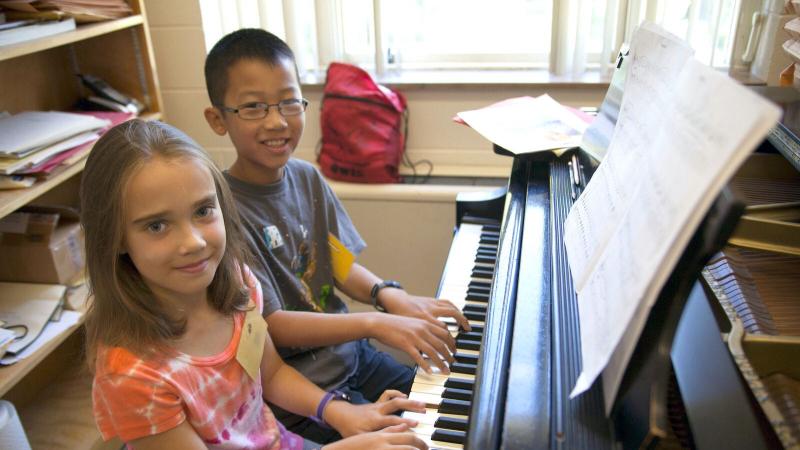 Two young students smile and sit at a piano keyboard in a music studio.