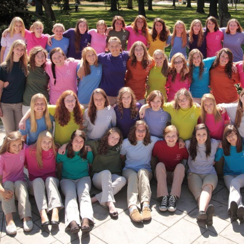 Bel Canto singers wearing Girl Choir t-shirts of all colors smile and pose with their arms around each other in the park, 
