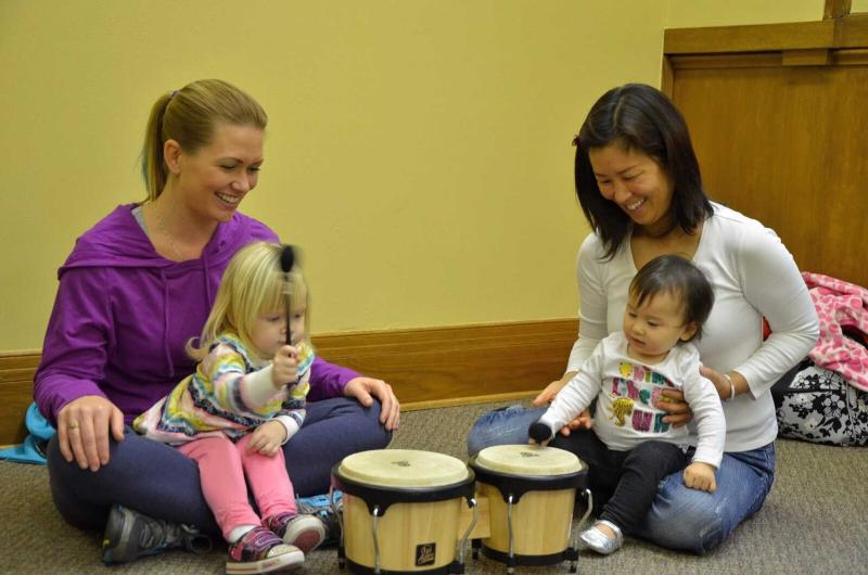 Two smiling parents and sit on the floor with toddlers on their laps in an Early Childhood Music class; the children hit a drum with mallets.