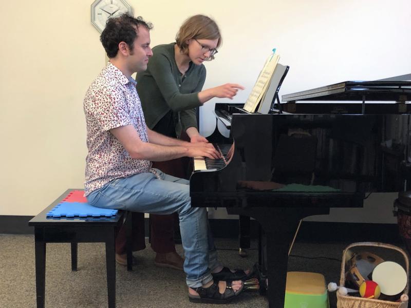 An adult student sits at a piano with his instructor correcting his hand posture and pointing at the score.