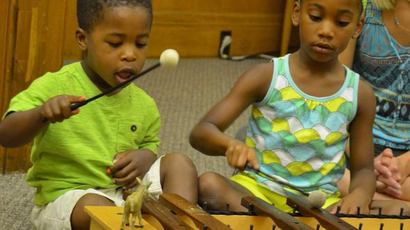 Two young siblings sit together in an Early Childhood Music class playing a xylophone togetherwith mallets.