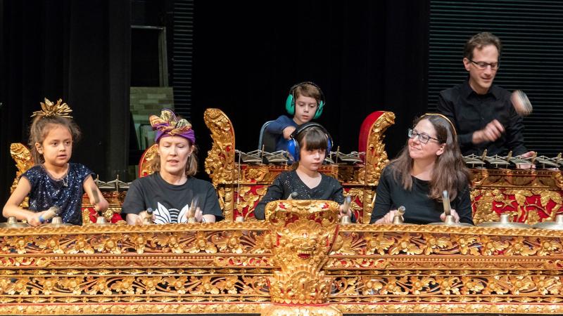 Children and adults play ornate, golden percussion instruments onstage during a Balinese Gamelan concert.
