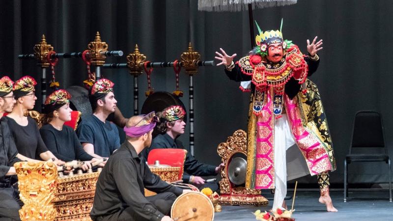 Students play ornate golden percussion instruments onstage and watch a dancer in a traditional Balinese gamelan performance outfit.
