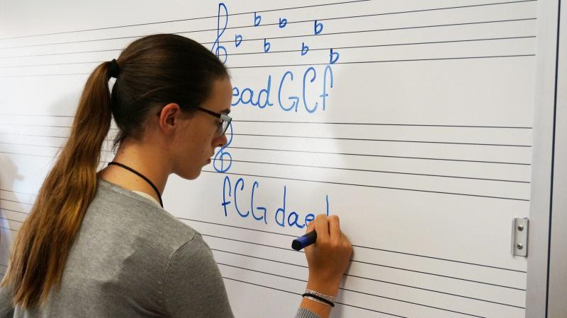 A student writes musical note names and symbols on a whiteboard.