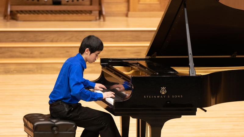 A student wearing a dress shirt and pants plays a grand piano on stage during the LCMS Honors Recital.