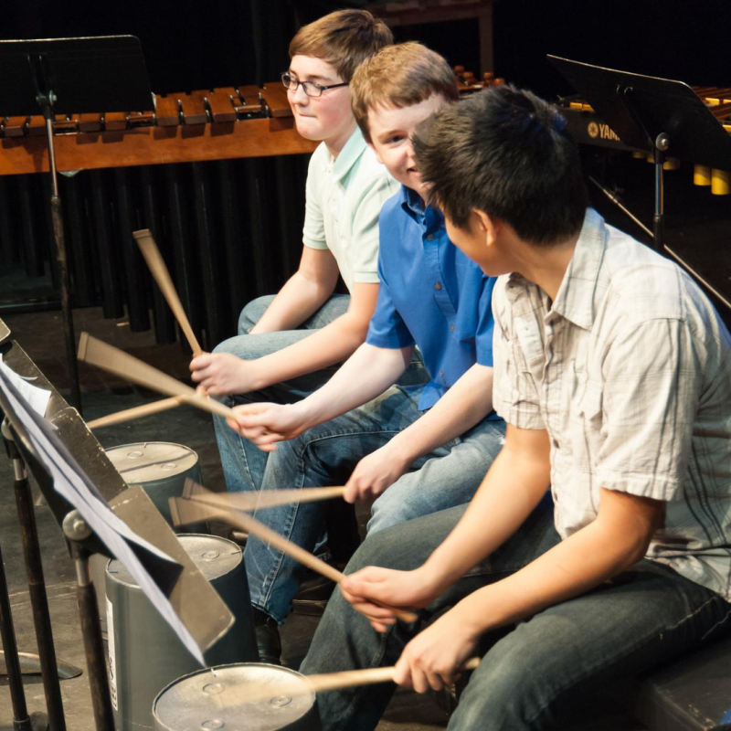 Three students drum with drumsticks and smile at each other, music stands in front of them.
