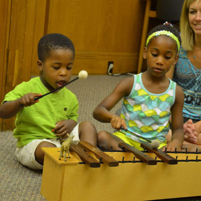 Two young siblings sit on the floor of an Early Childhood Music class and play a xylophone with mallets.
