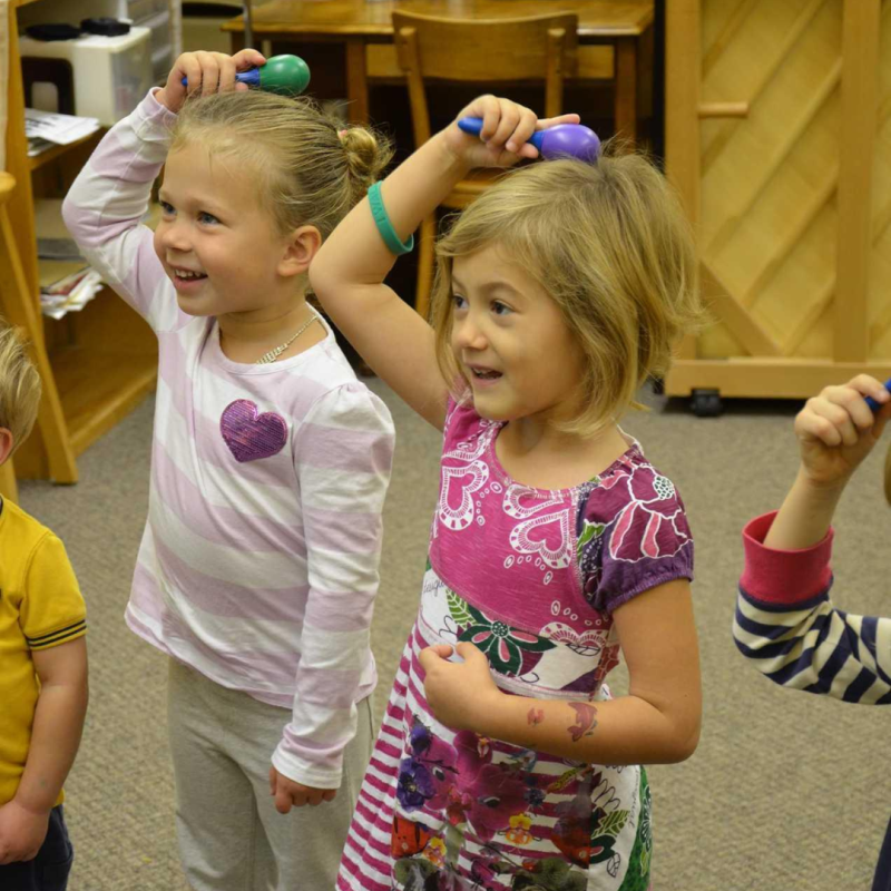 Pre-K children stand together in an early Childhood Music class, smiling and patting their heads with shakers.