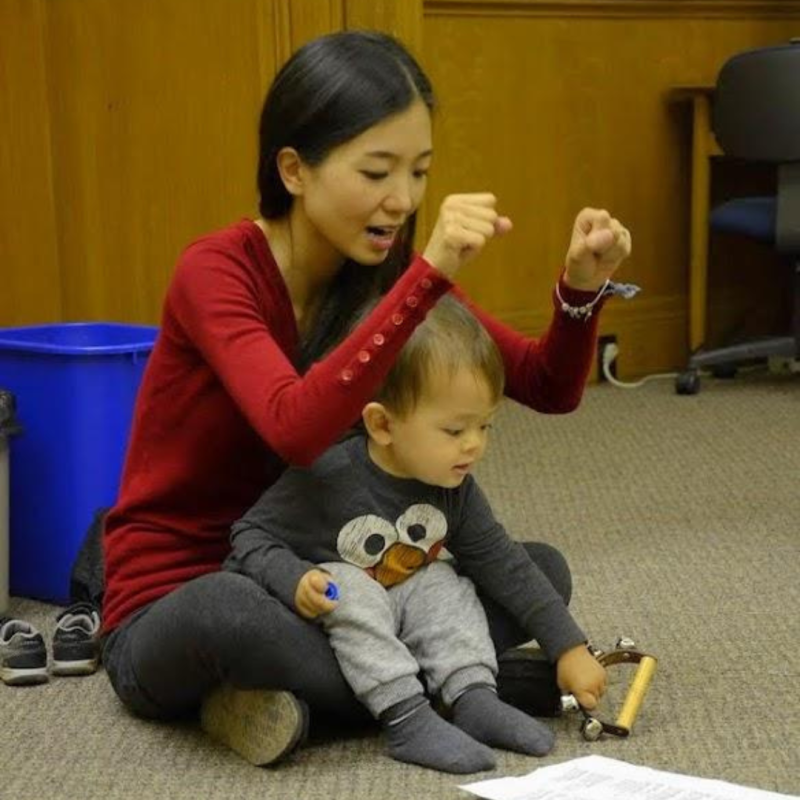 A mother sits on the floor with her child in her lap. She sings and motions with her hands while the toddler waves handbells.