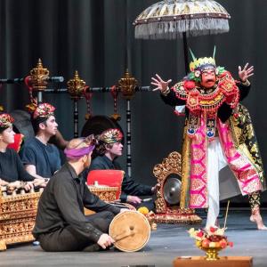 Students play ornate golden percussion instruments onstage and watch a dancer in a traditional Balinese gamelan performance outfit.
